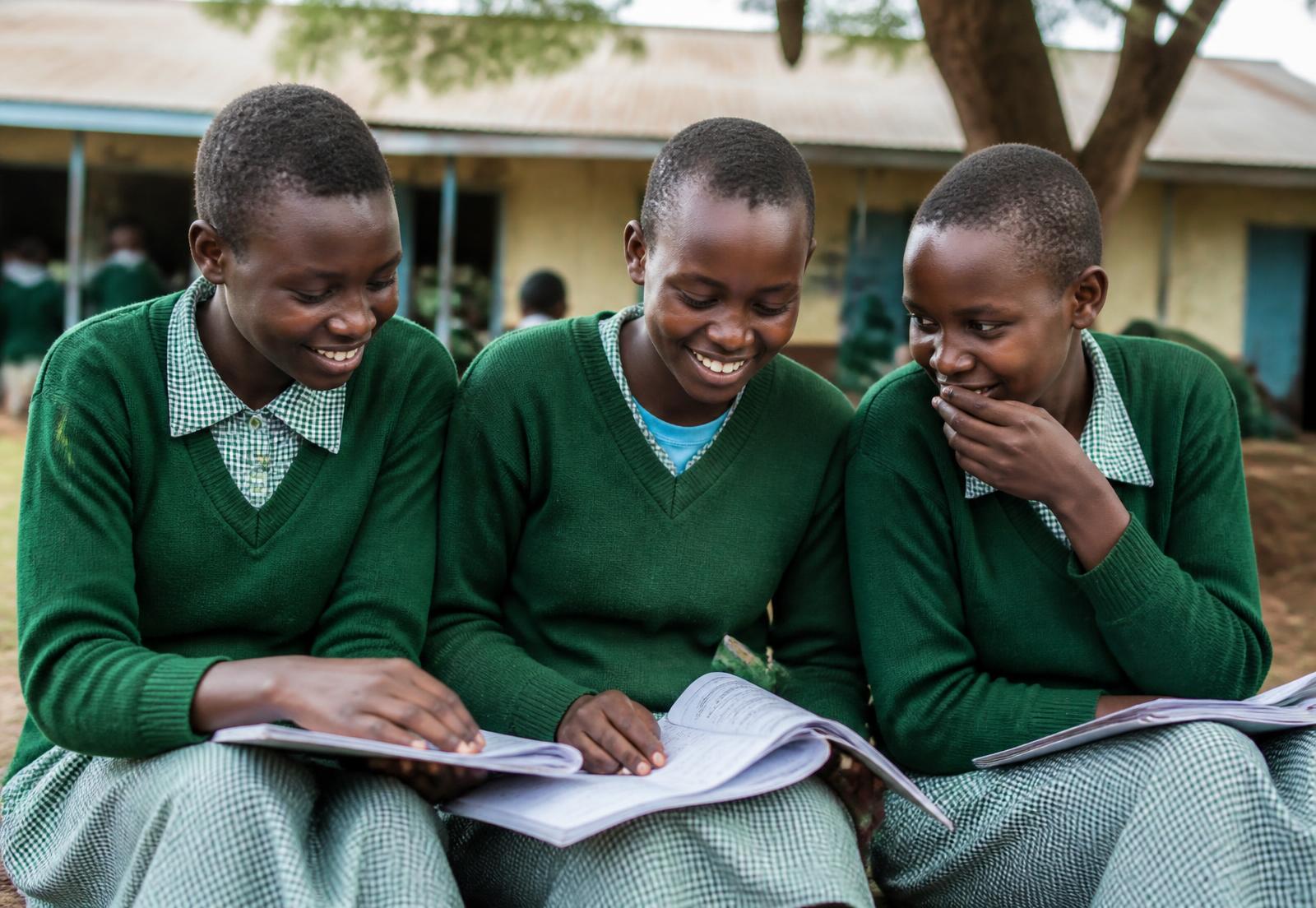 Three Kenyan schoolgirls in green uniforms reading together outside their classroom